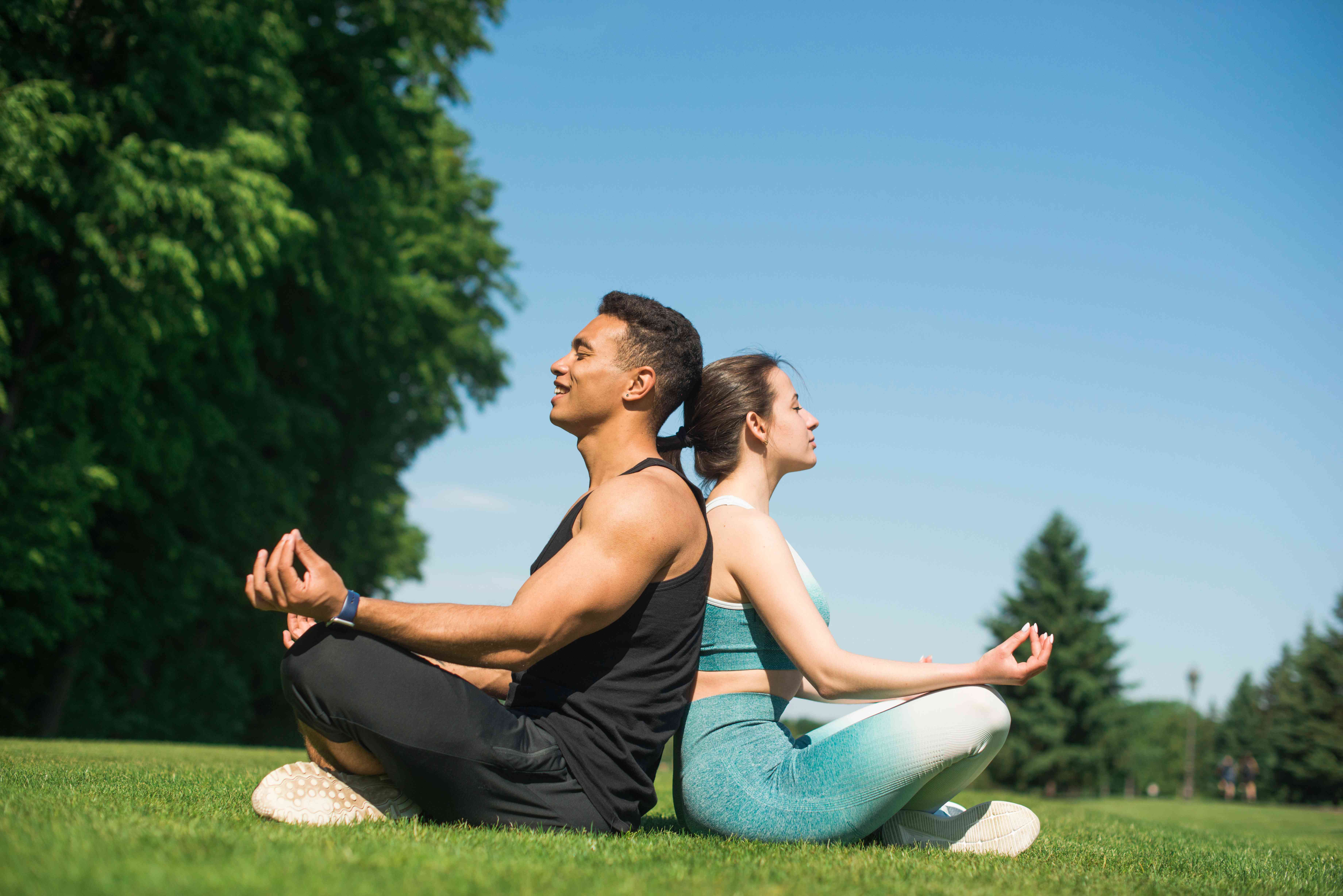 Pareja joven practicando yoga y meditación al aire libre en un parque - Hombre y mujer en posición de loto espalda con espalda, representando bienestar, salud integral, vida saludable y equilibrio físico-mental que promueve ALDIMED